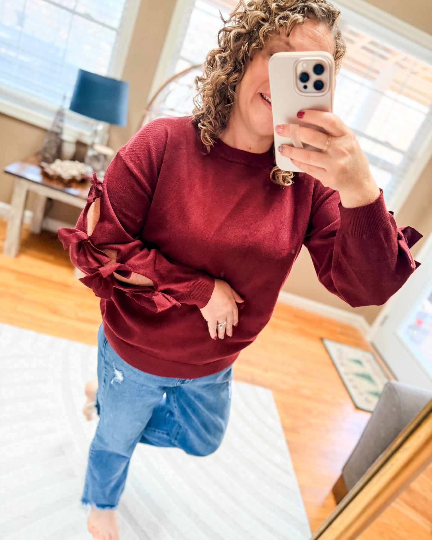 Person taking a mirror selfie wearing a red sweater and blue jeans in a room with a desk and computer.