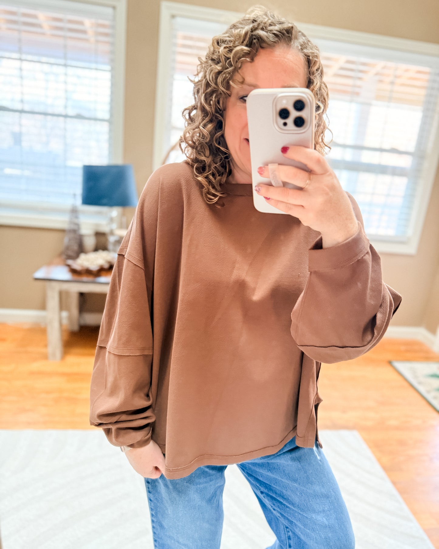 Person wearing a brown oversized sweatshirt and blue jeans taking a mirror selfie.