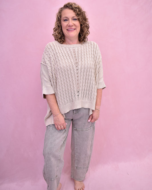 Woman wearing a cream knitted sweater and light gray pants against a pink background