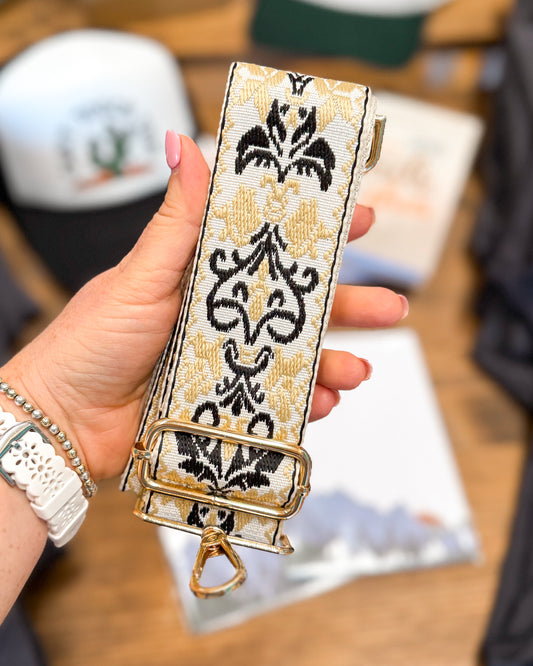 Hand holding a decorative strap with intricate black and gold patterns on a blurred background