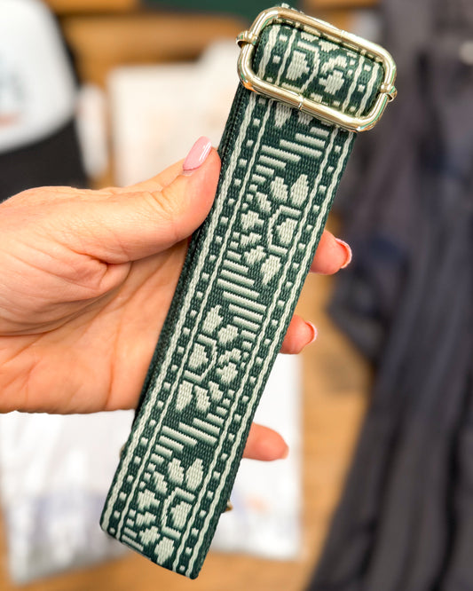 Hand holding a green textured strap with a metal buckle against a blurred background