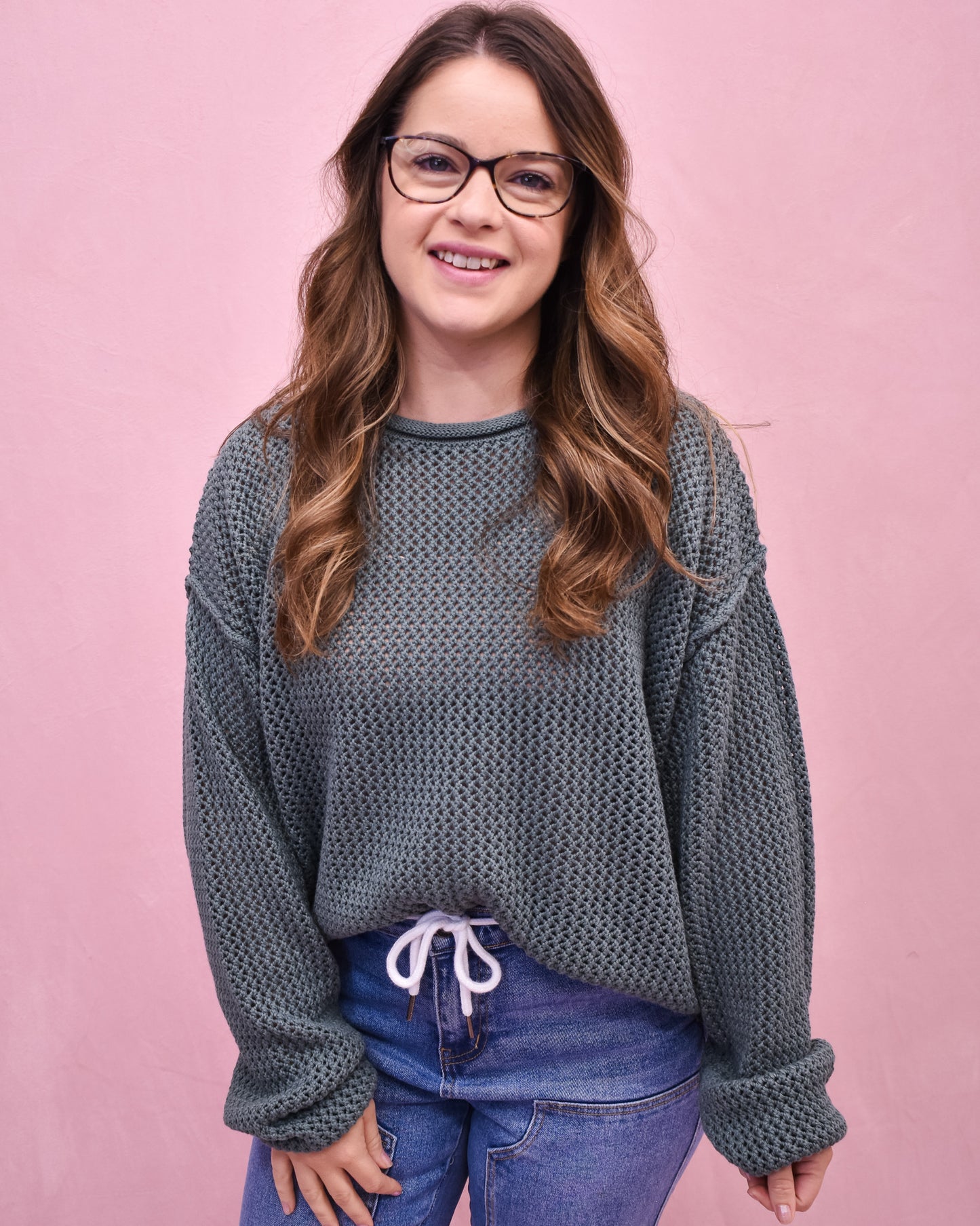 Woman wearing a textured gray sweater and blue jeans against a pink background