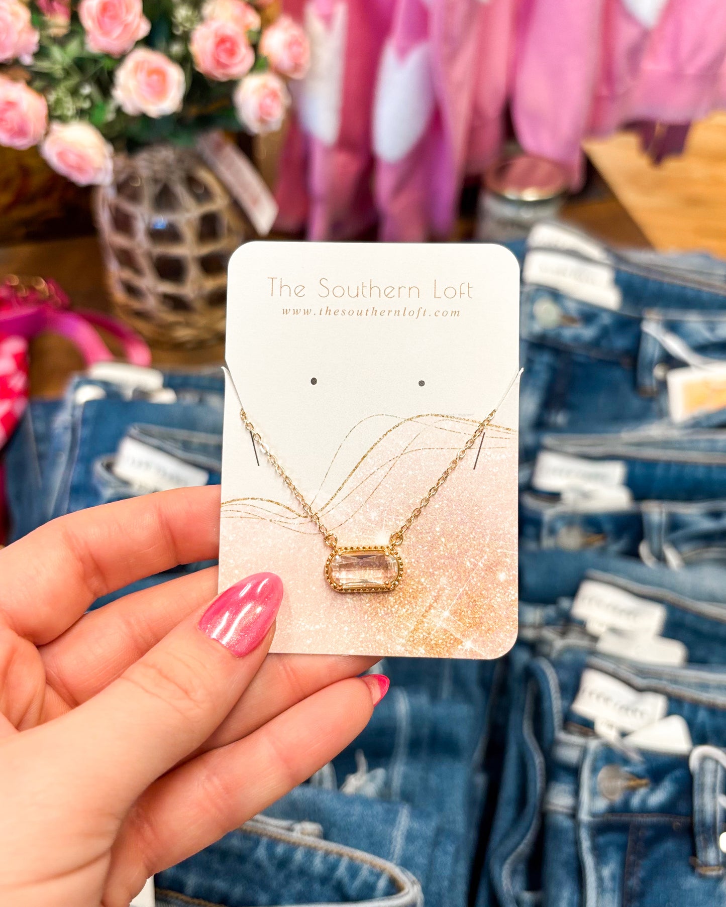 Gold necklace on a card held by a hand with pink nail polish, denim jeans in the background