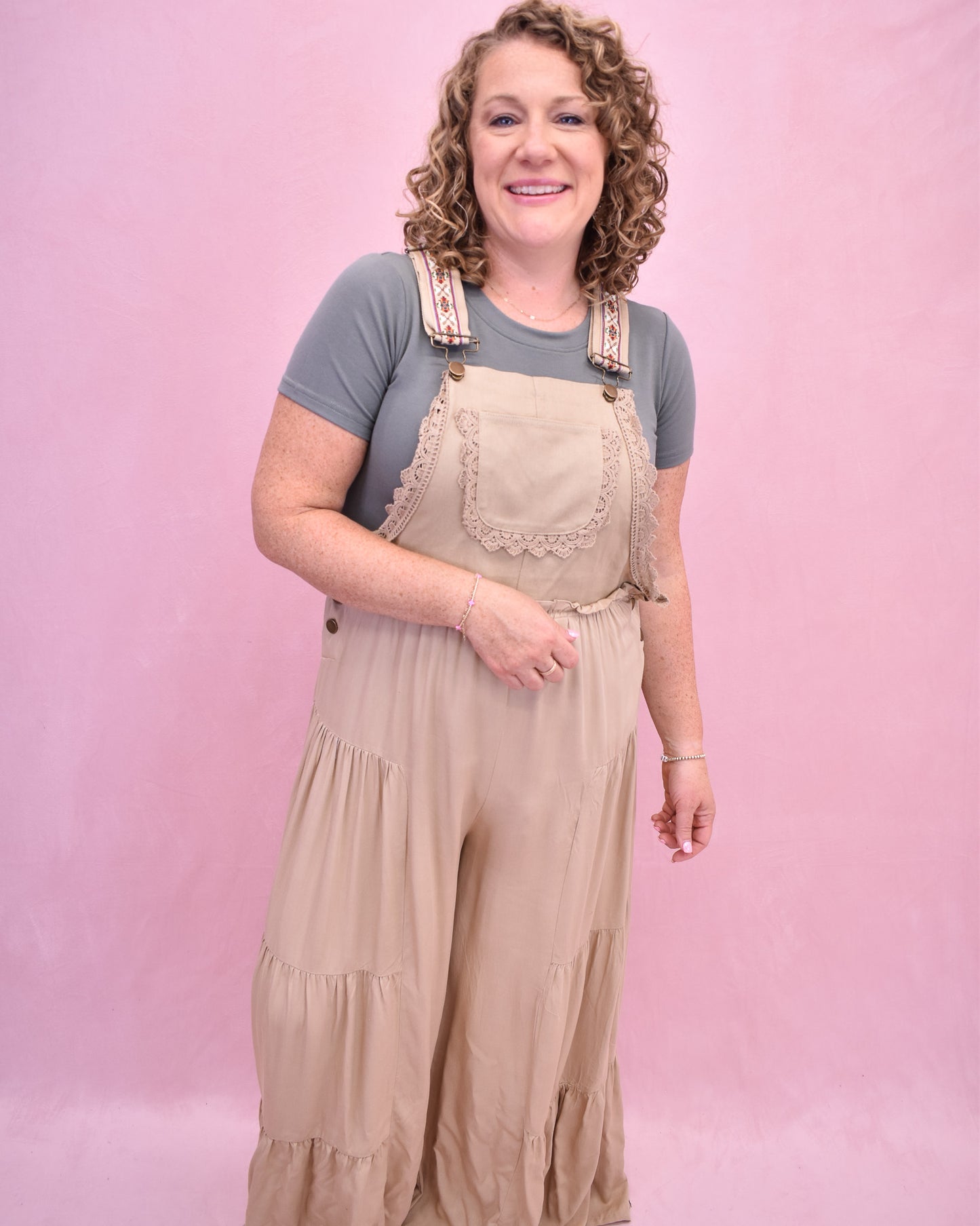 Woman wearing beige overalls and a gray shirt against a pink background