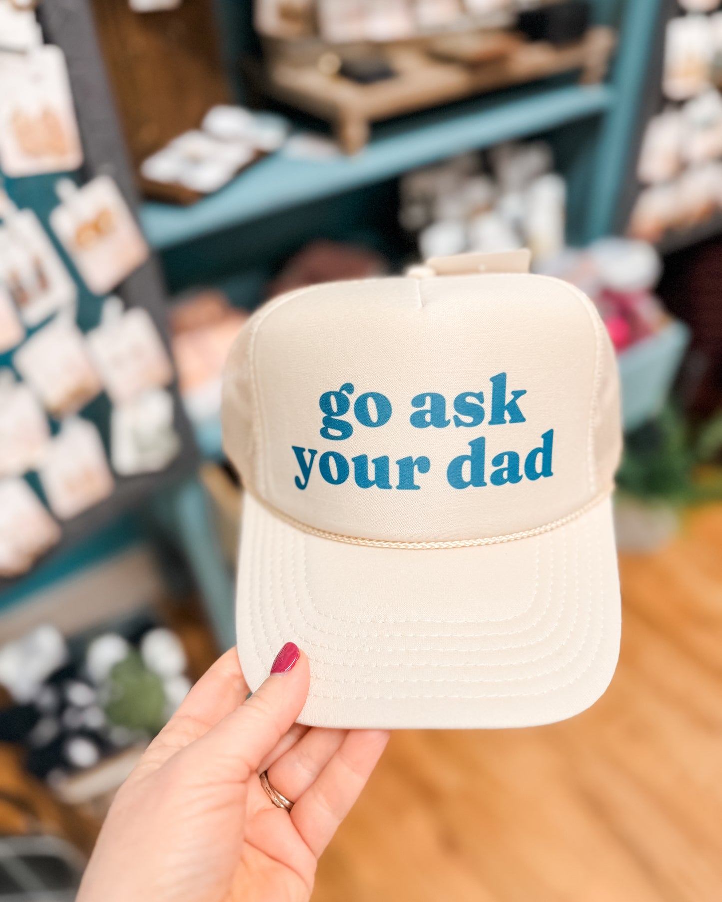 Beige cap with 'go ask your dad' text held in a hand with a store in the background