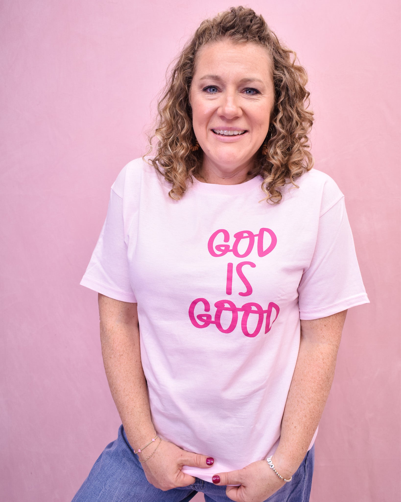 Woman wearing a white t-shirt with 'GOD IS GOOD' in pink text against a pink background
