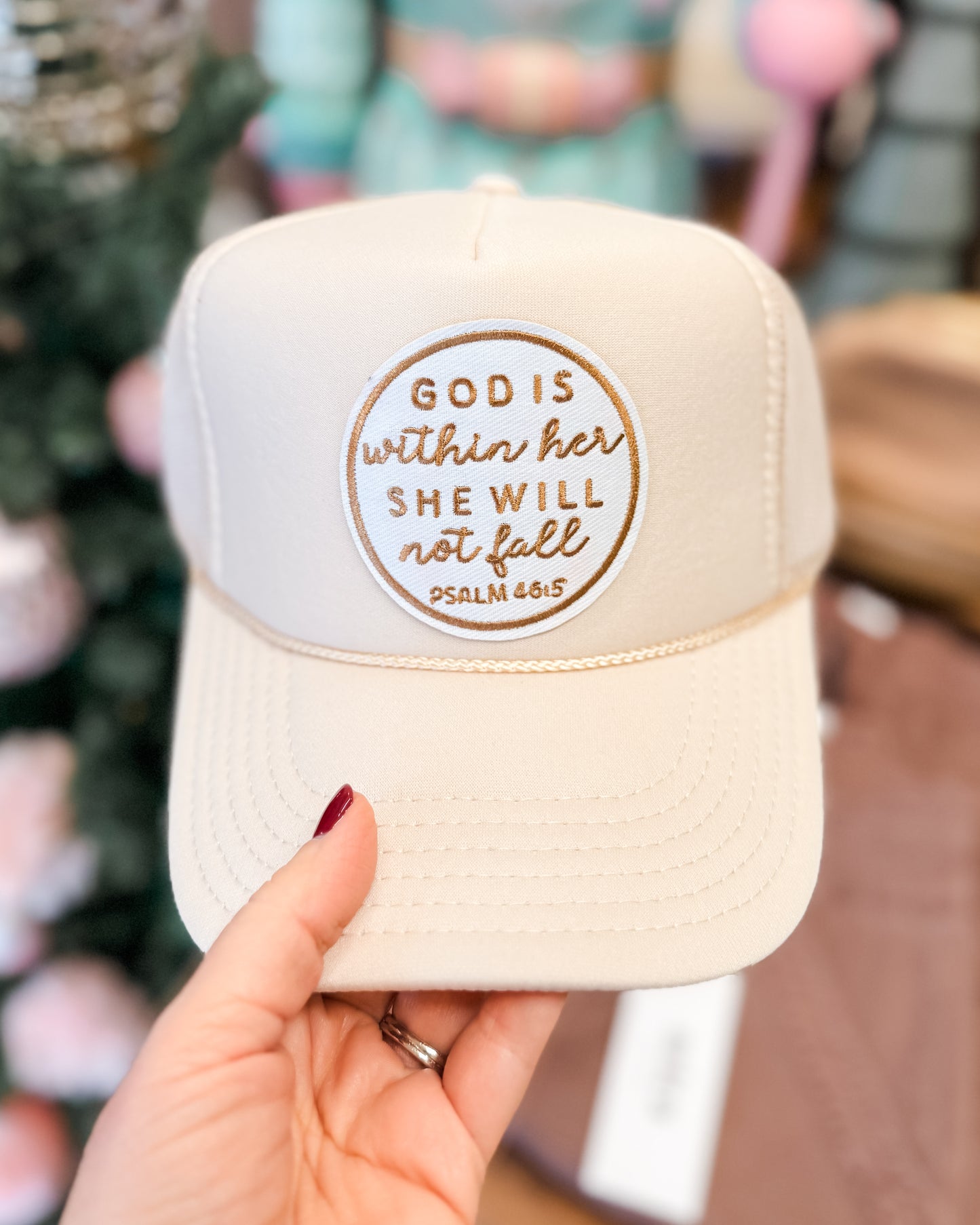 Beige cap with 'God is within her, she will not fall' quote held by a hand against a blurred background