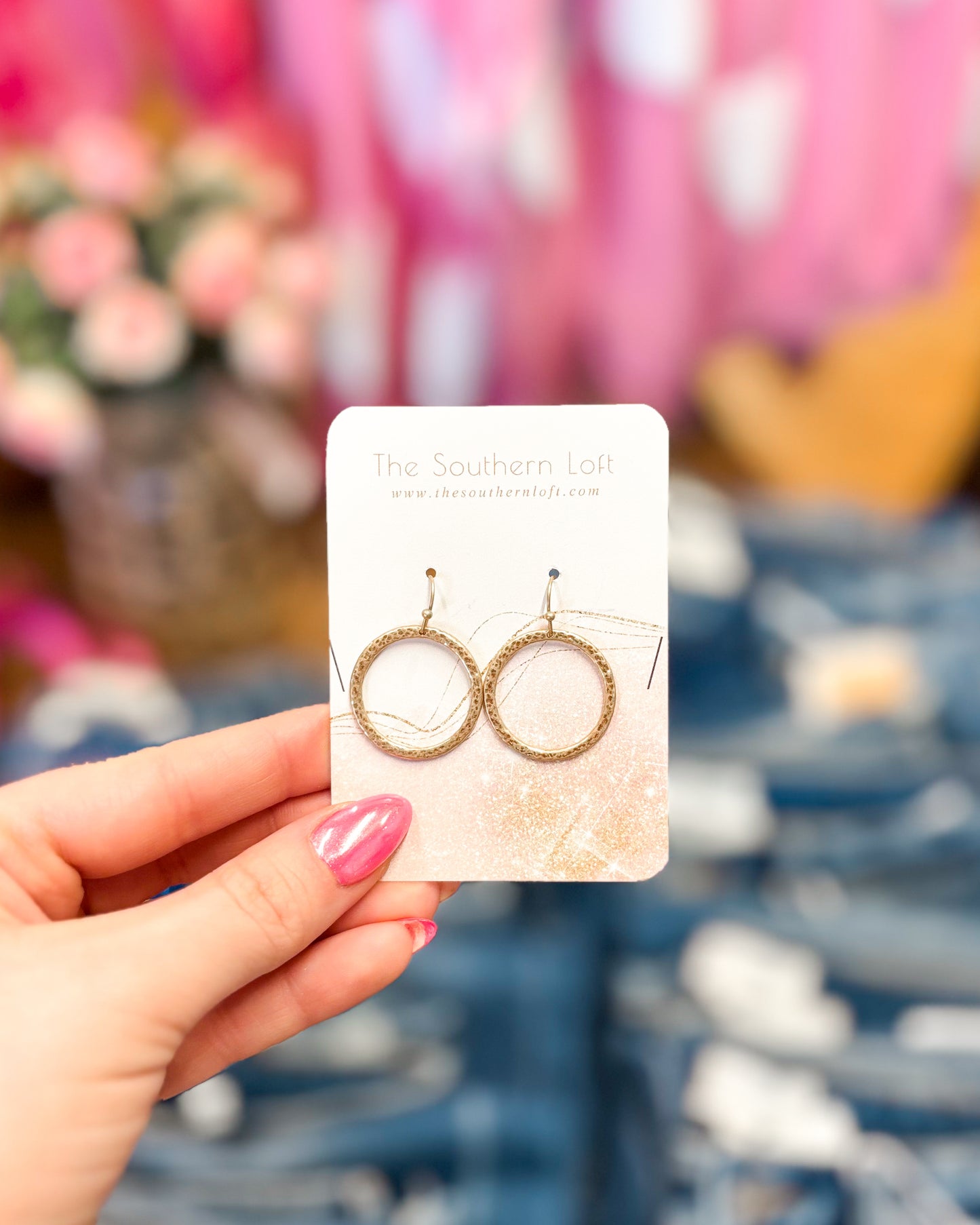 Gold hoop earrings on a card held by a hand with pink nail polish, blurred floral and fabric background