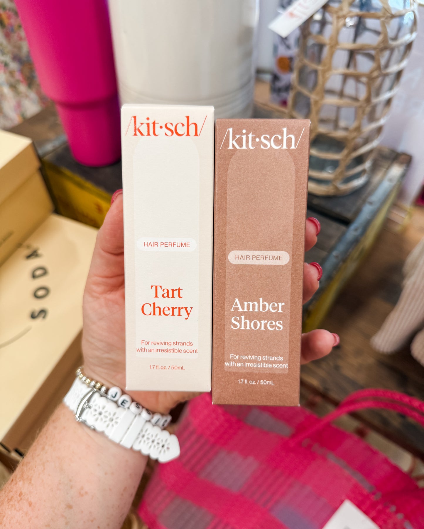 Two Kitsch hair perfume bottles, 'Tart Cherry' and 'Amber Shores', held in a hand with a blurred background.