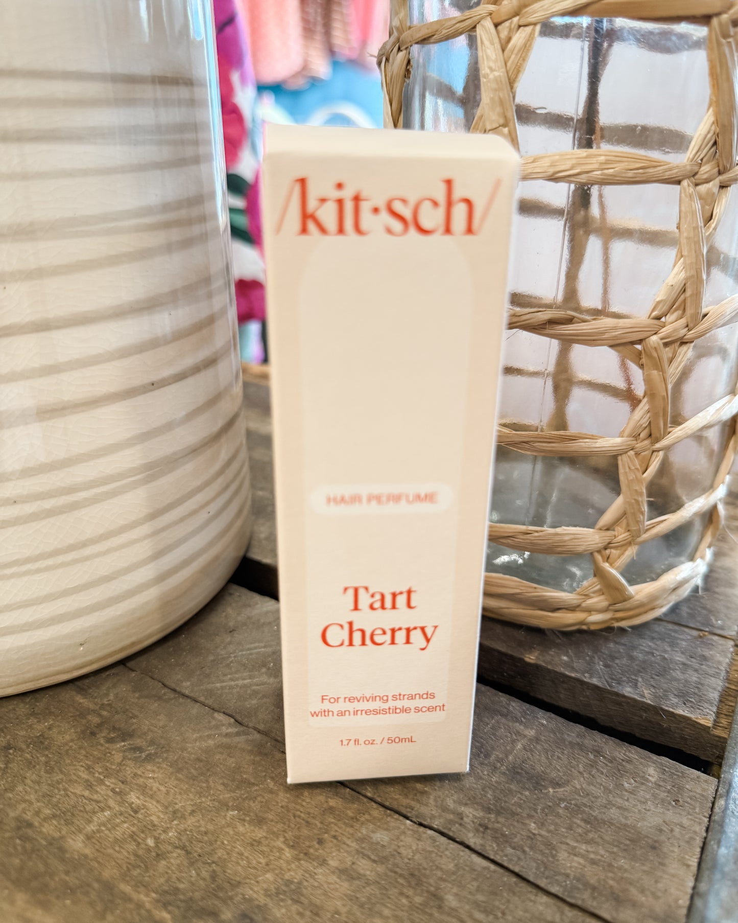 Kitsch Tart Cherry perfume bottle on a wooden surface with decorative vases in the background