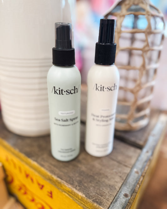 Two white spray bottles with black nozzles on a wooden surface, featuring the 'kitsch' brand.
