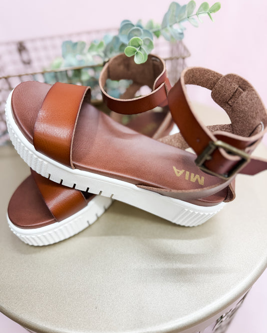 Brown sandals with white soles on a light surface with a plant in the background