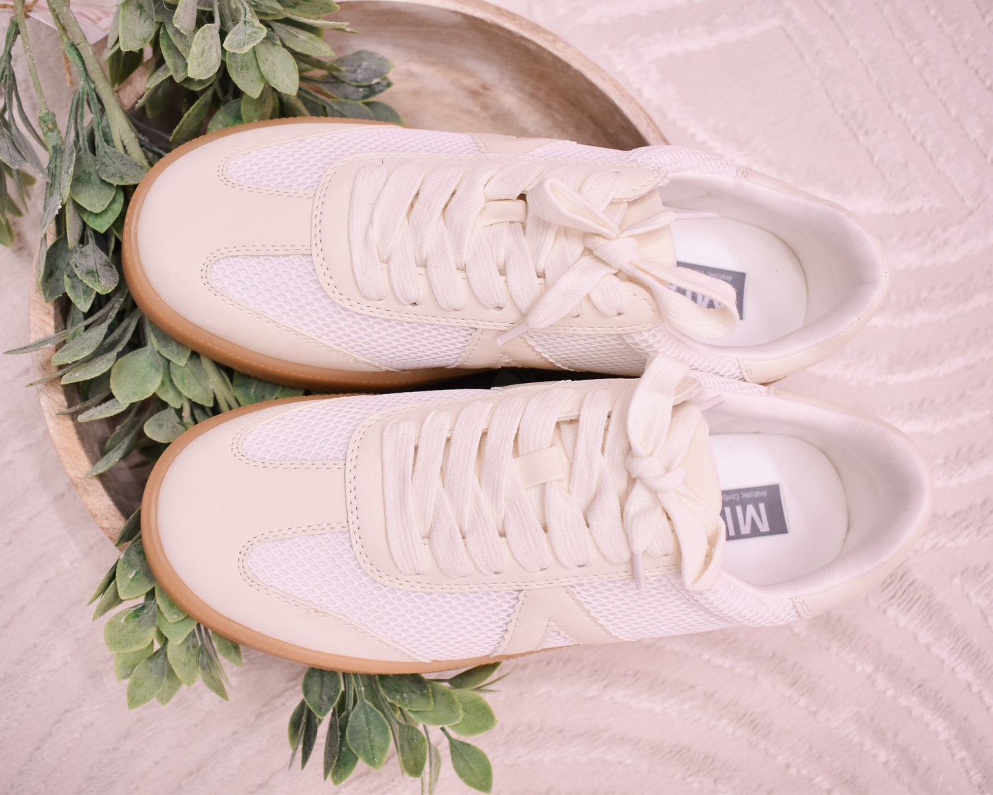 Pair of white sneakers with brown soles on a textured surface with green leaves.