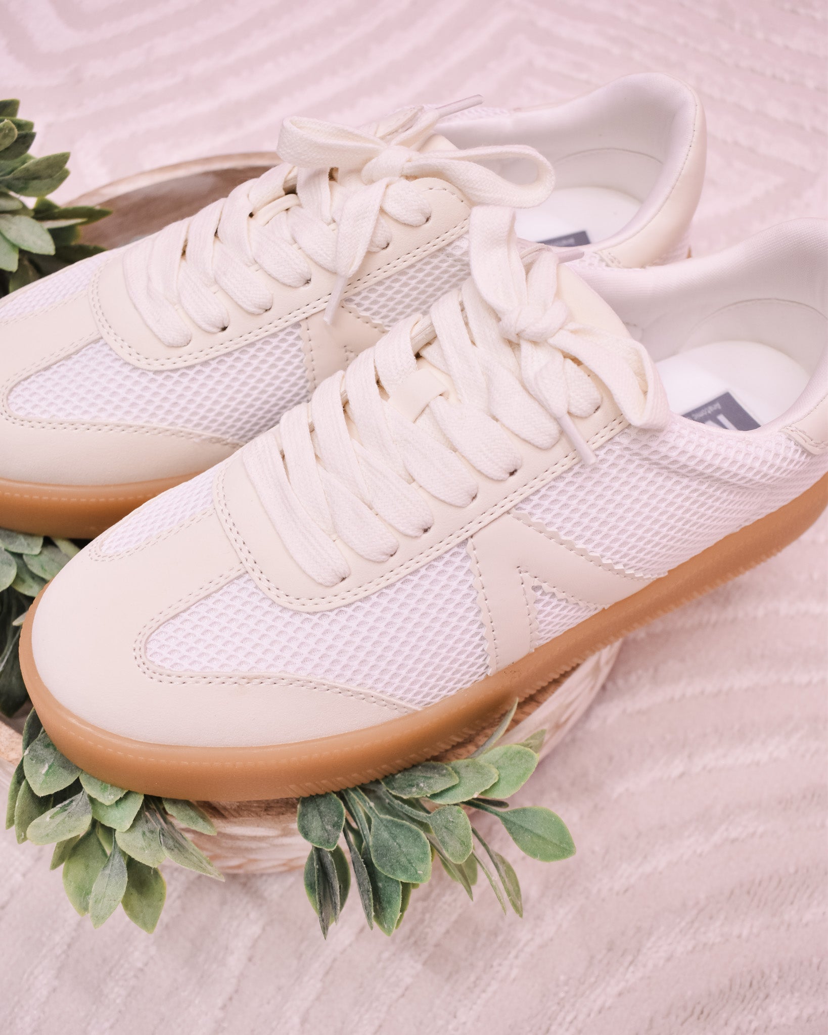 White sneakers with brown soles on a light pink surface with green leaves.