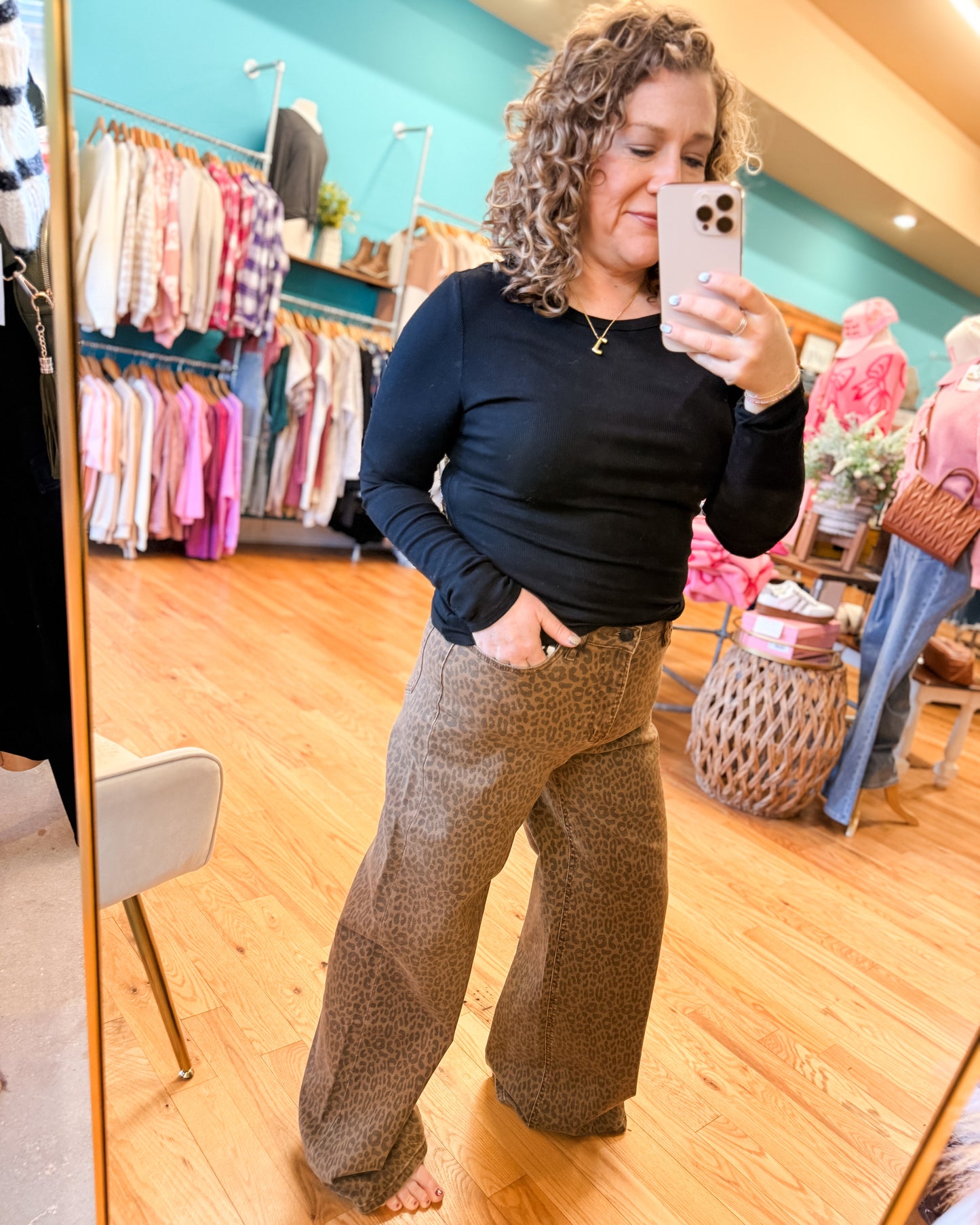 Woman taking a mirror selfie in a clothing store wearing a black long-sleeve top and brown patterned pants.