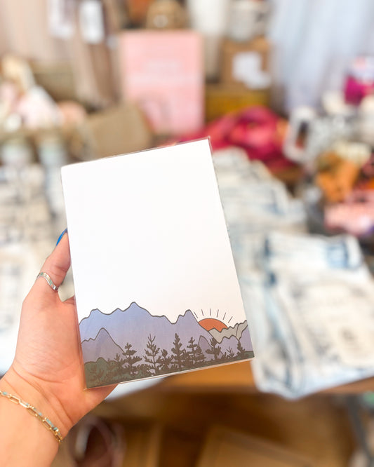 Hand holding a postcard with a mountain and sun design against a blurred indoor background