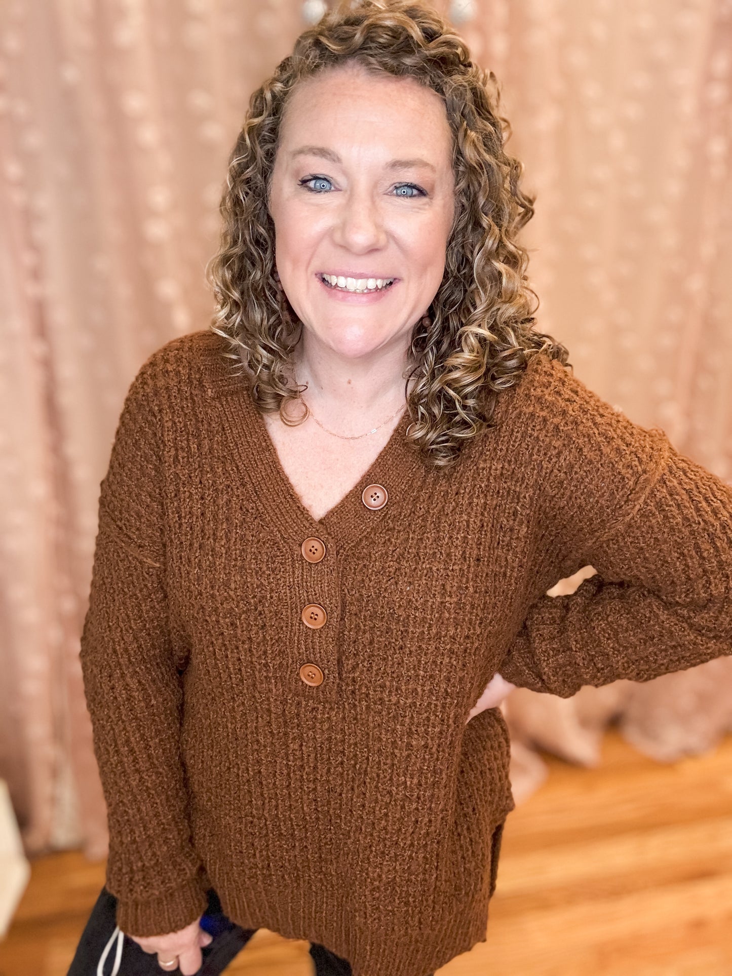A smiling woman wearing a brown V-neck sweater with button trim details.