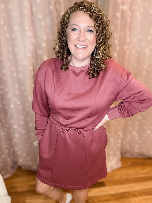 A person standing in a room with a pink mini dress featuring a crew neckline and elastic waist with front tie.