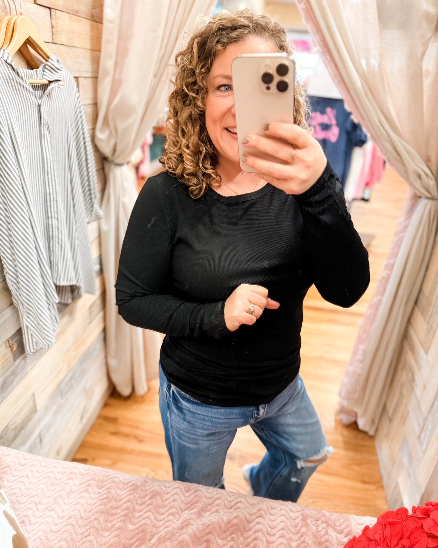 Woman taking a mirror selfie wearing a black long-sleeve top and blue jeans in a clothing store.