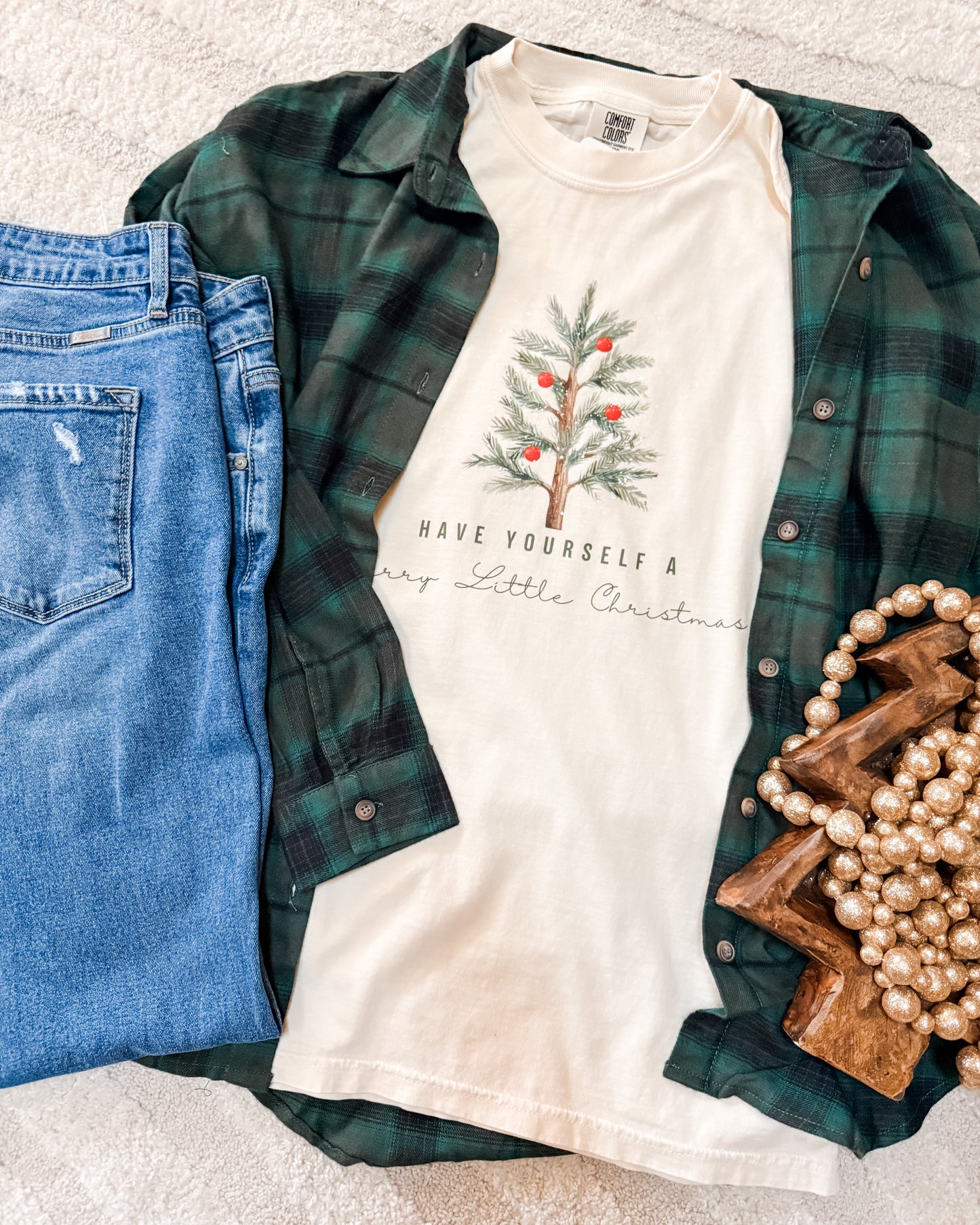 White t-shirt with Christmas tree design and text, green plaid shirt, blue jeans, and decorative items on a light gray surface.