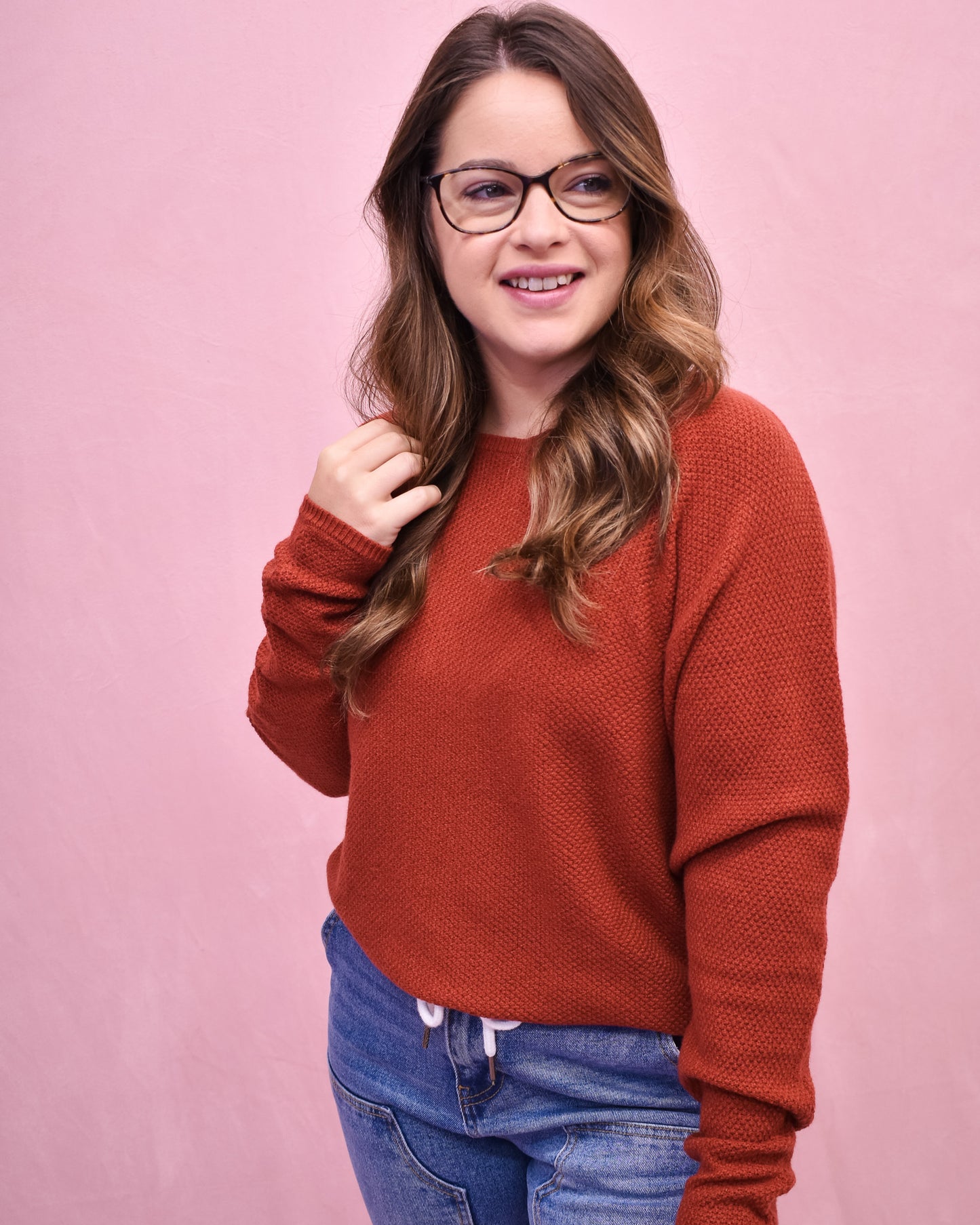 Woman wearing a rust-colored sweater and blue jeans against a pink background