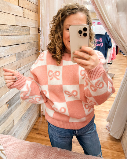 Person wearing a pink and white checkered sweater with bow patterns, taking a mirror selfie.