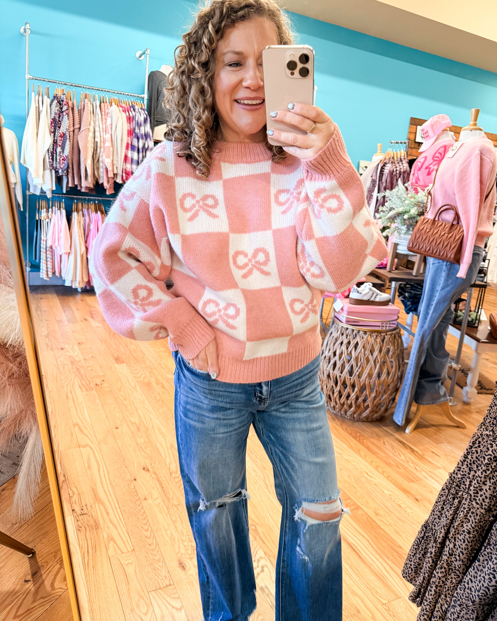Woman taking a mirror selfie wearing a pink and white checkered sweater with bow designs and blue jeans in a clothing store.