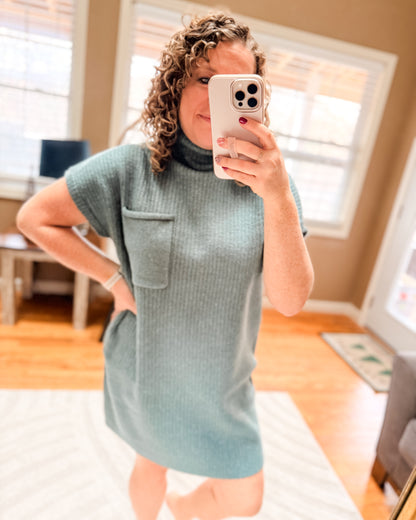 Woman taking a mirror selfie wearing a light blue dress in a room with wooden flooring and windows.
