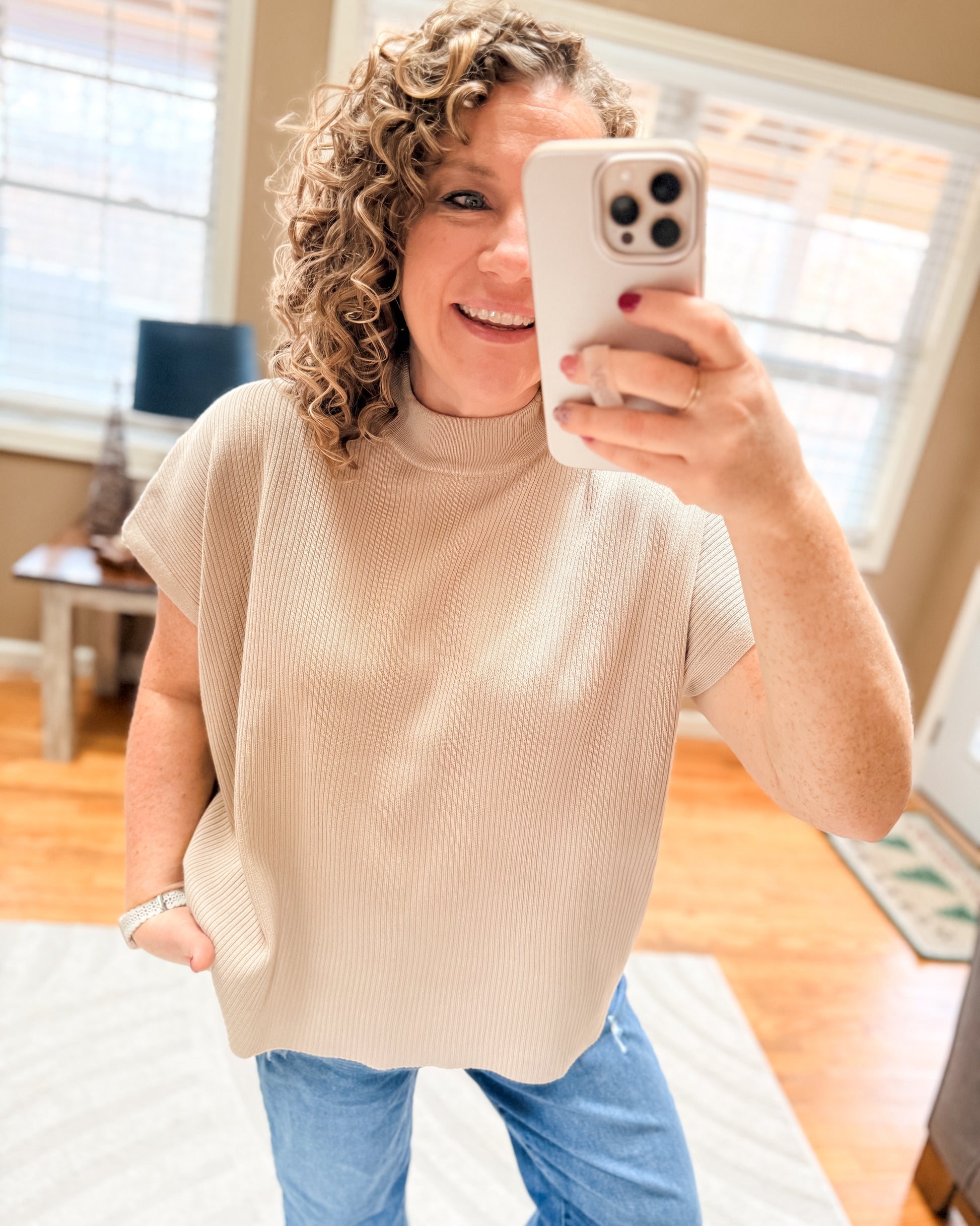 Woman taking a mirror selfie wearing a beige sleeveless sweater and blue jeans indoors.