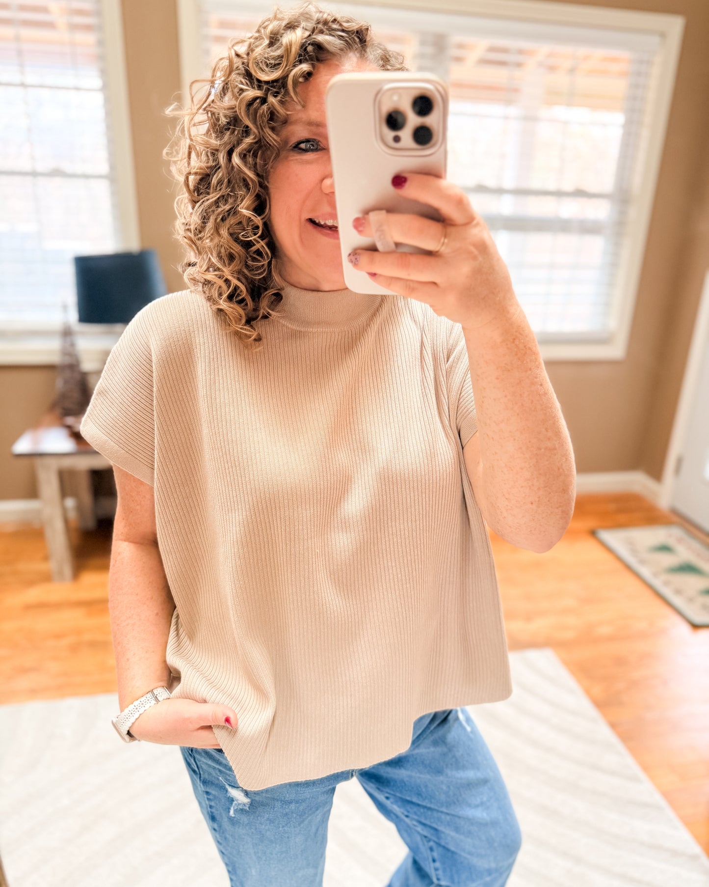 Woman taking a mirror selfie wearing a beige top and blue jeans in a room with wooden flooring.