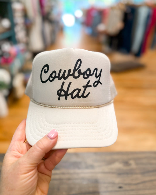 Beige cap with 'Cowboy Hat' text held by a hand in a store setting