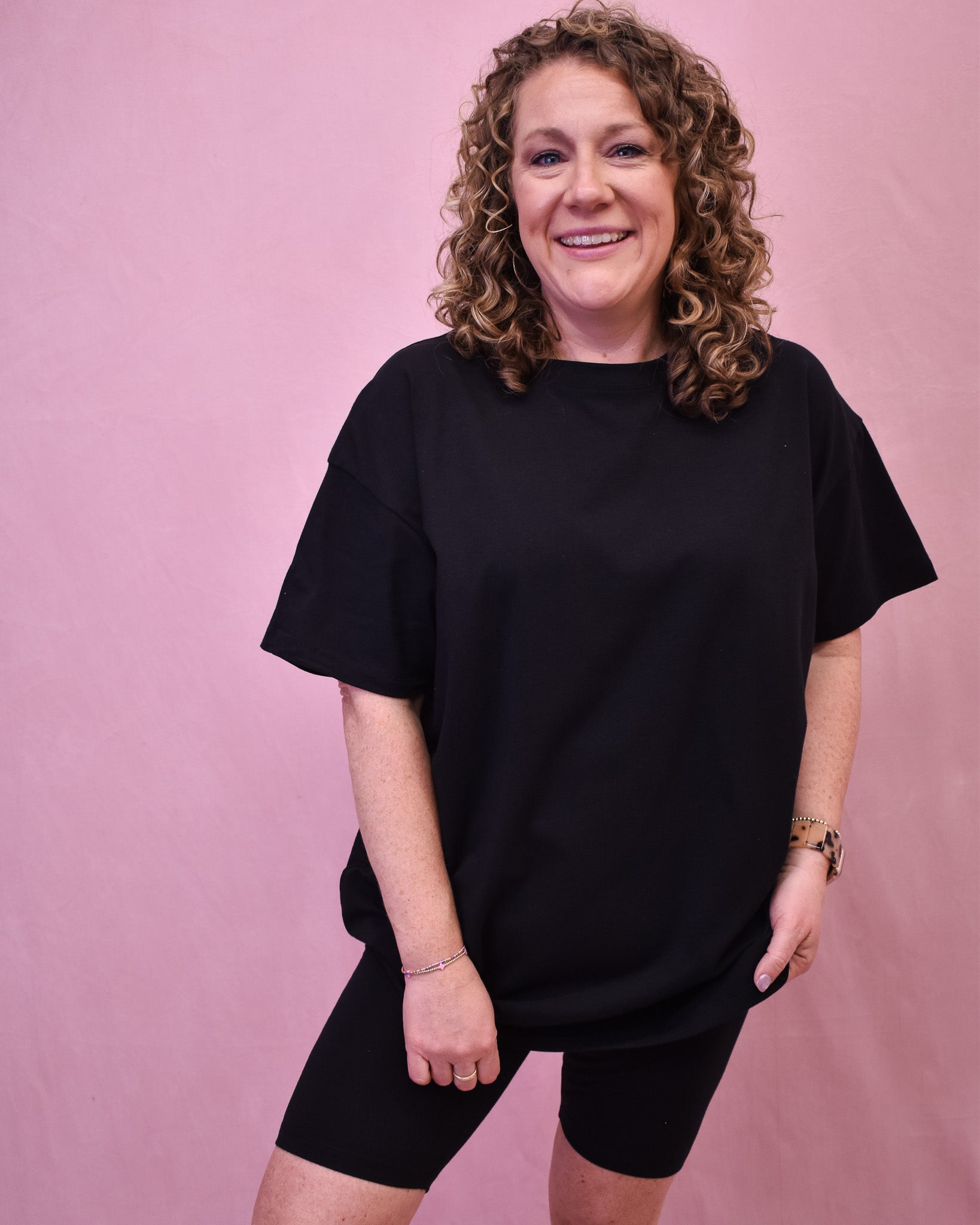 Person wearing a black oversized t-shirt and black shorts against a pink background