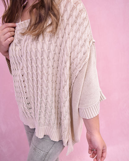 Person wearing a beige cable knit sweater against a pink background
