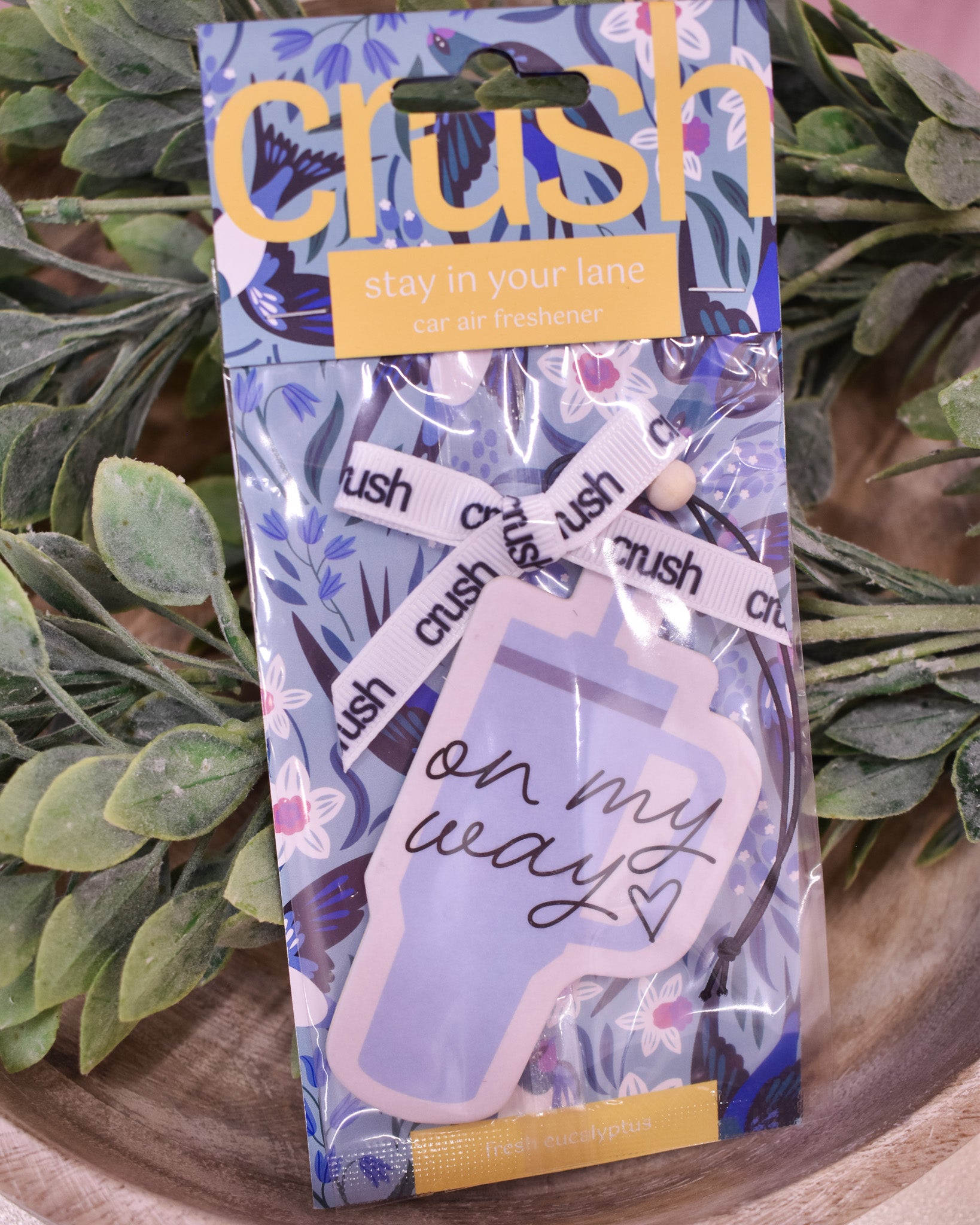 Packaged 'Crush' car air freshener with decorative elements on a wooden surface with plants in the background.
