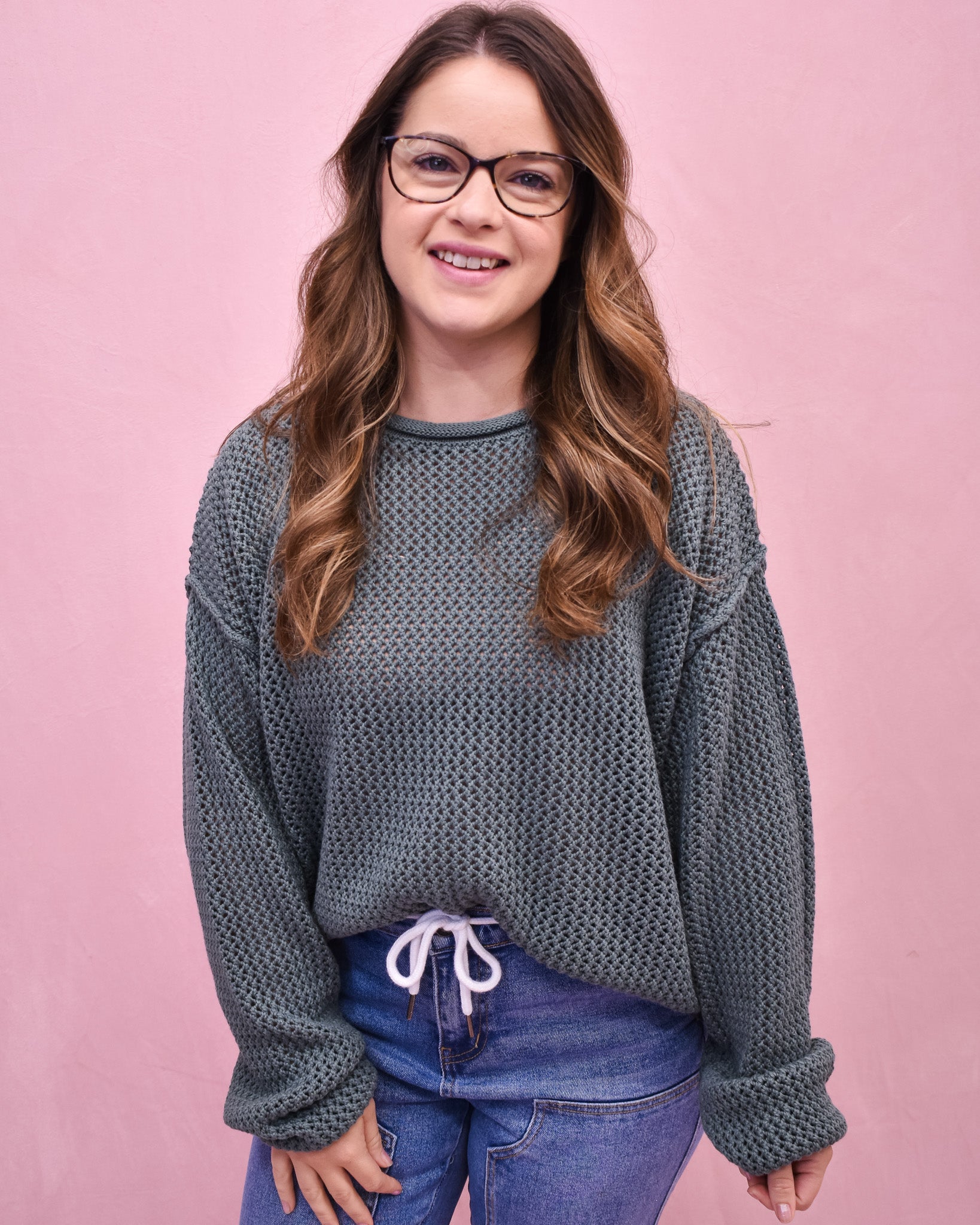 Woman wearing a textured gray sweater and blue jeans against a pink background