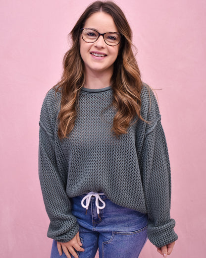 Woman wearing a textured gray sweater and blue jeans against a pink background