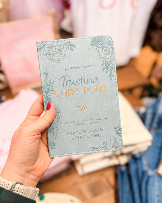 Hand holding a book titled 'Trusting God's Plan' with floral design on the cover.