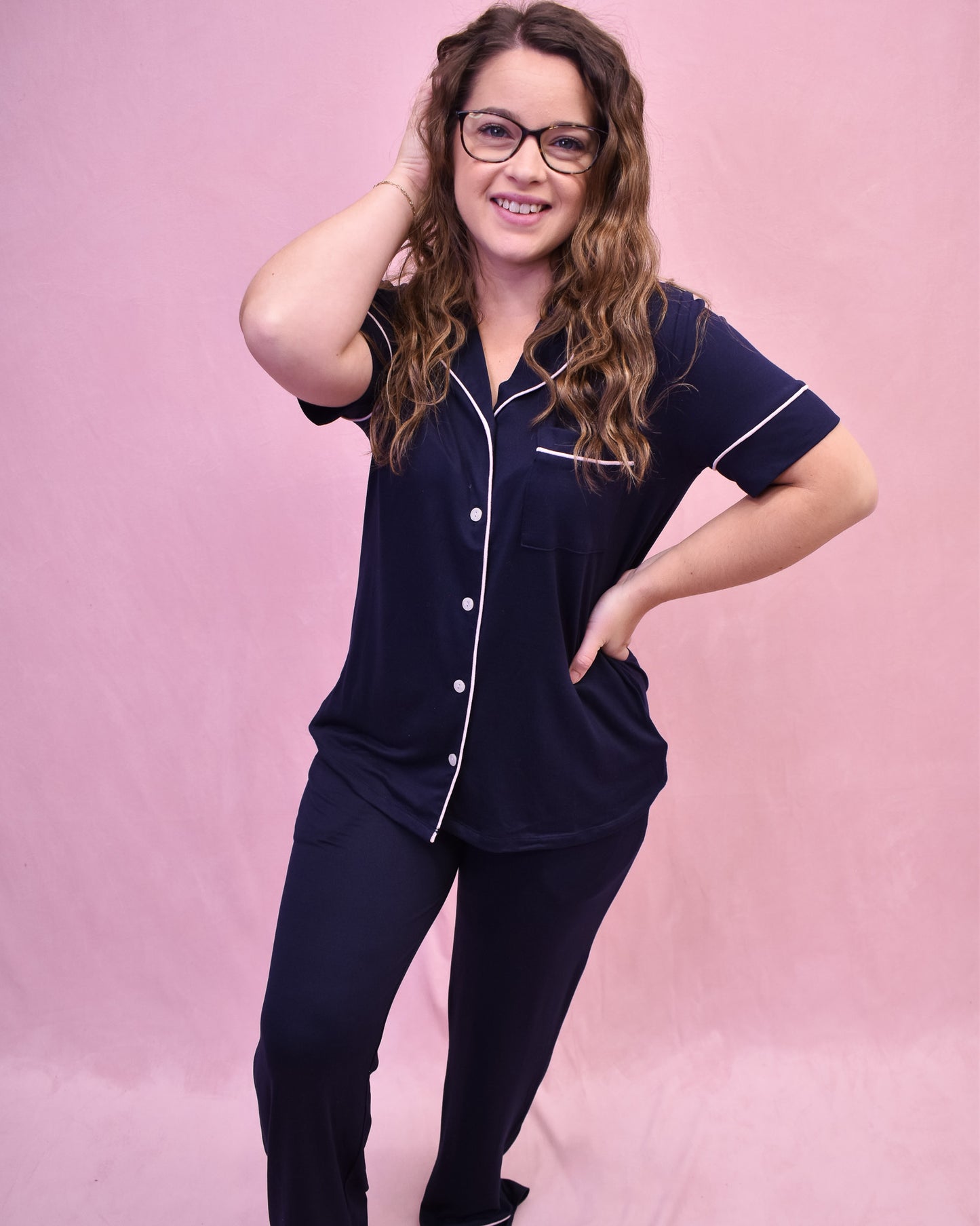 Woman wearing a navy pajama set with white piping against a pink background