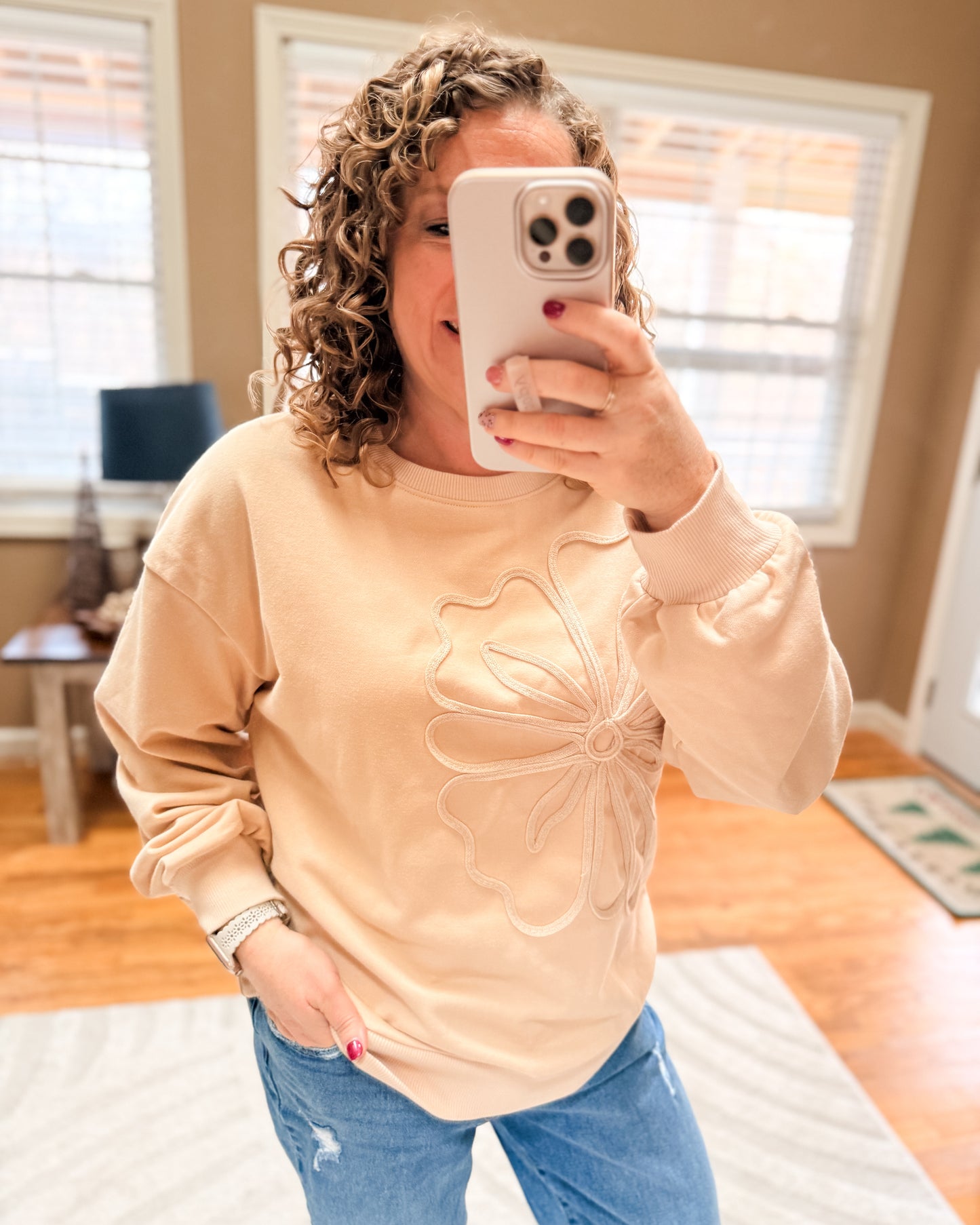 Person wearing a beige sweater with a floral design in a room with wooden flooring and a lamp.