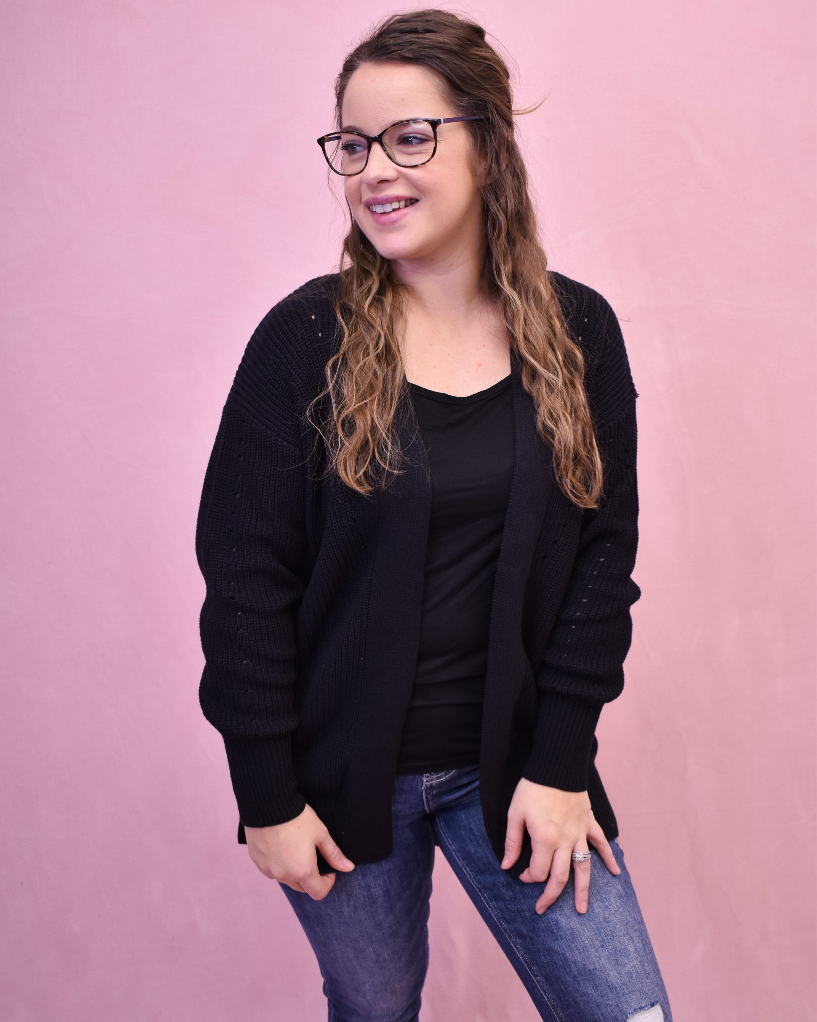 Woman wearing a black cardigan over a black top and blue jeans against a pink background