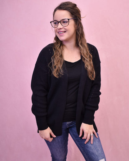 Woman wearing a black cardigan over a black top and blue jeans against a pink background