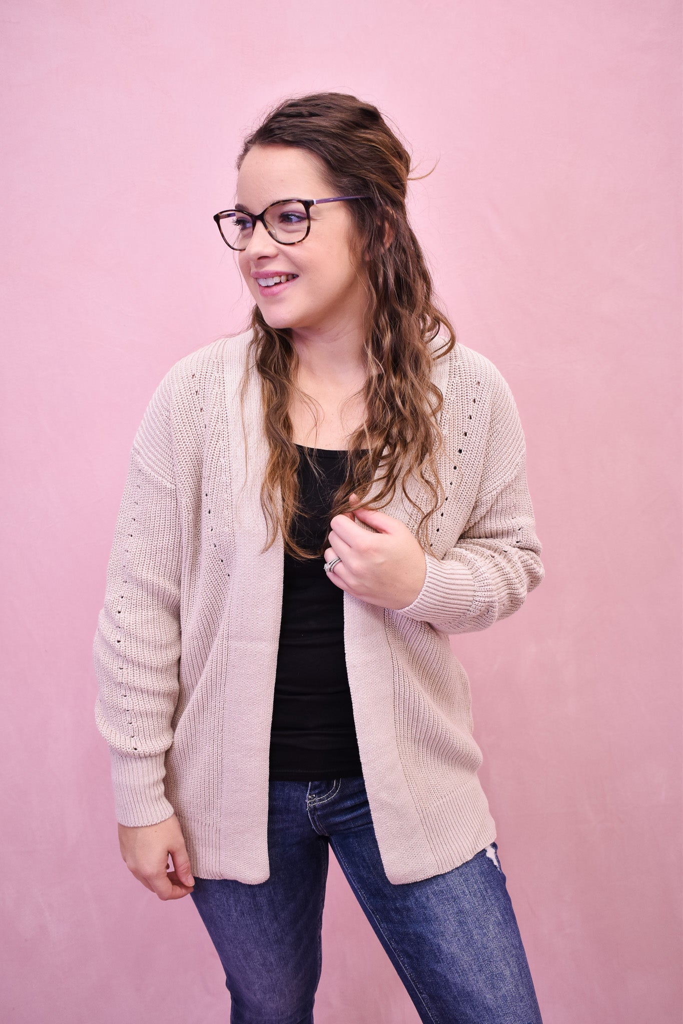 Woman wearing a light pink cardigan over a black top and blue jeans on a pink background
