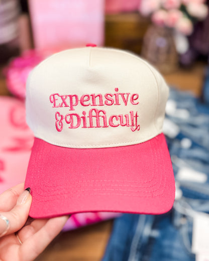 Pink and white cap with 'Expensive & Difficult' text held by a hand.