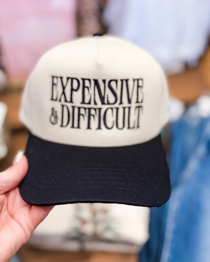 Cap with 'EXPENSIVE & DIFFICULT' text held by a hand against a blurred background