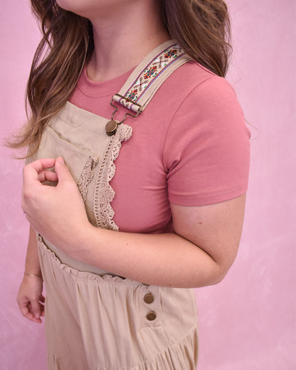 Person wearing a pink shirt and beige overalls with floral straps against a pink background