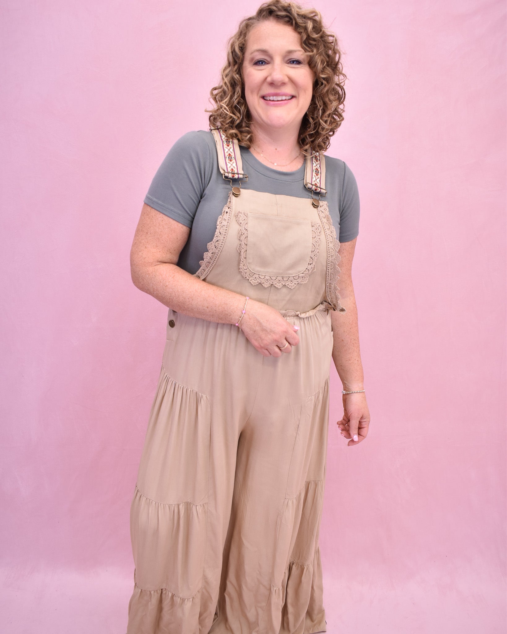 Woman wearing beige overalls and a gray shirt against a pink background