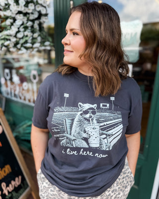a short sleeve graphic top i live here now with a racoon at a football field holding an iced coffee on a dark gray tee.