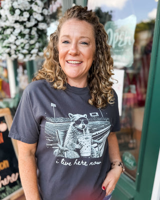 a short sleeve graphic top i live here now with a racoon at a football field holding an iced coffee on a dark gray tee.