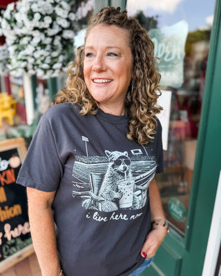 a short sleeve graphic top i live here now with a racoon at a football field holding an iced coffee on a dark gray tee.