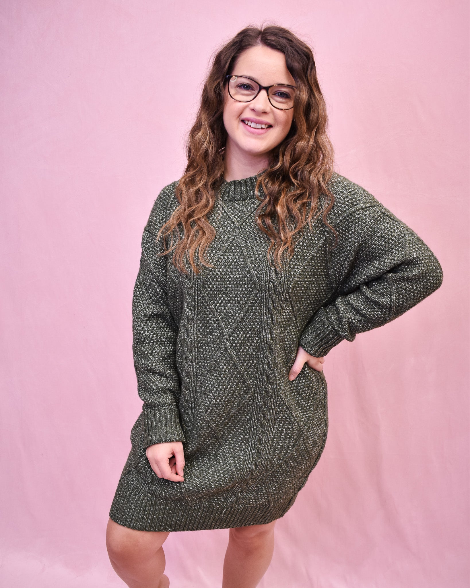 Woman wearing a green cable knit sweater dress against a pink background